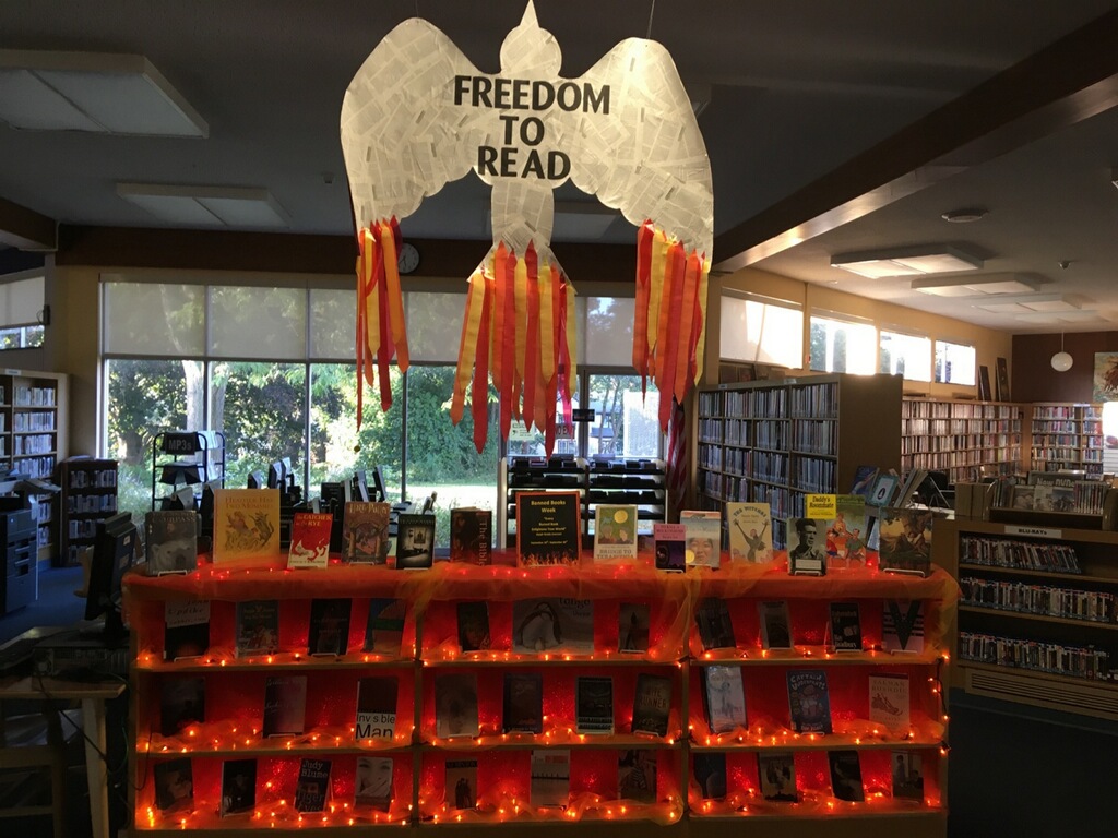 An outline of a bird wallpapered with book pages hangs above a shelf of books. The shelves have orange tealights strung across them.