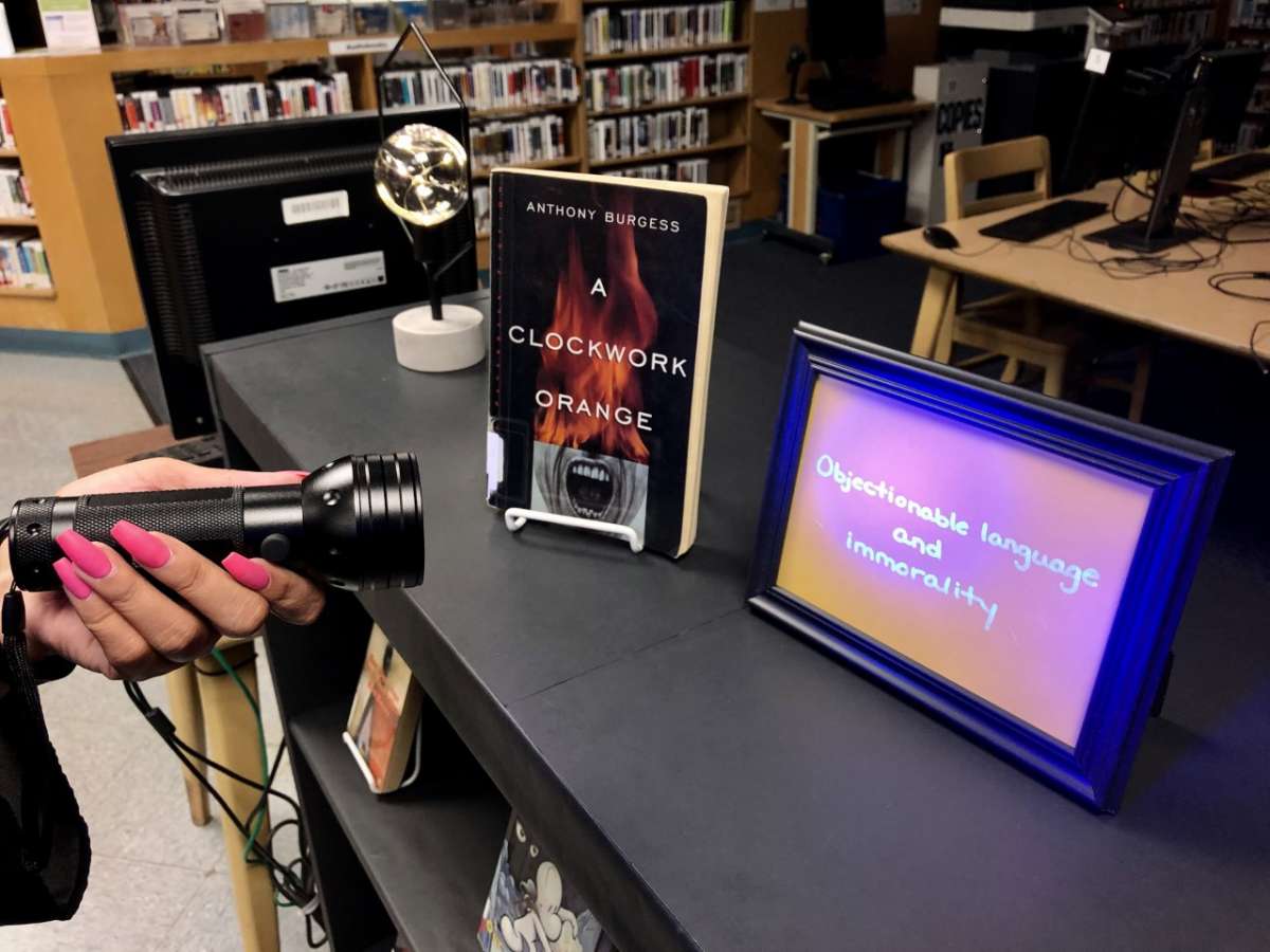 a person shines a black light flashlight at a frame next to A Clockwork Orange, illuminating the reasons why the book was challenged on the frame