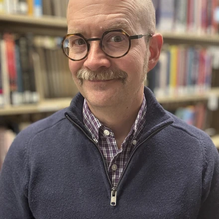 Image of a bald white man with a mustache and glasses in front of books