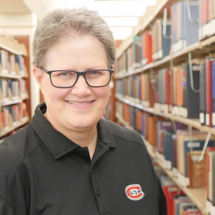 woman in glasses wearing a black polo shirt. she is standing in front of library bookshelves