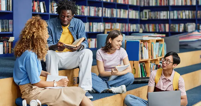 A group of four students talking and studying in a library with bookshelves in the background.