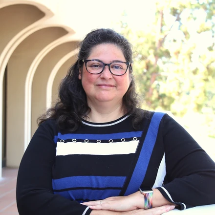 Natalie Marquez with brown hair and glasses, wearing blue, black, and white sweater standing outside UC Irvine, Langson Library.